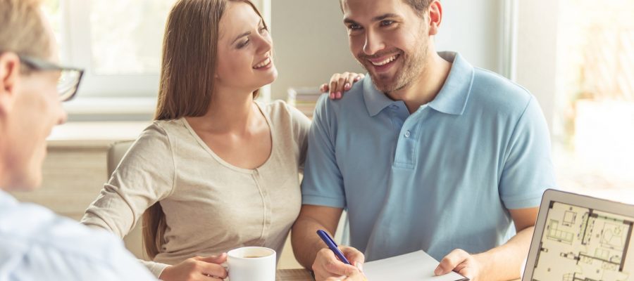 Handsome middle aged realtor in classic shirt and eyeglasses and happy young couple are discussing new house. Couple is drinking coffee, making notes and smiling while sitting in office
